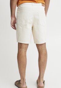 Pantalones cortos de algodón beige con bolsillos traseros, trabillas para el cinturón y un corte recto. Llevados con zapatos marrones de tipo slip-on. Textura y diseño simples.