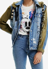 Chaqueta de denim con un acabado azul claro desgastado, mangas de color oliva y una camiseta gráfica que presenta un colorido diseño de Mickey Mouse debajo.