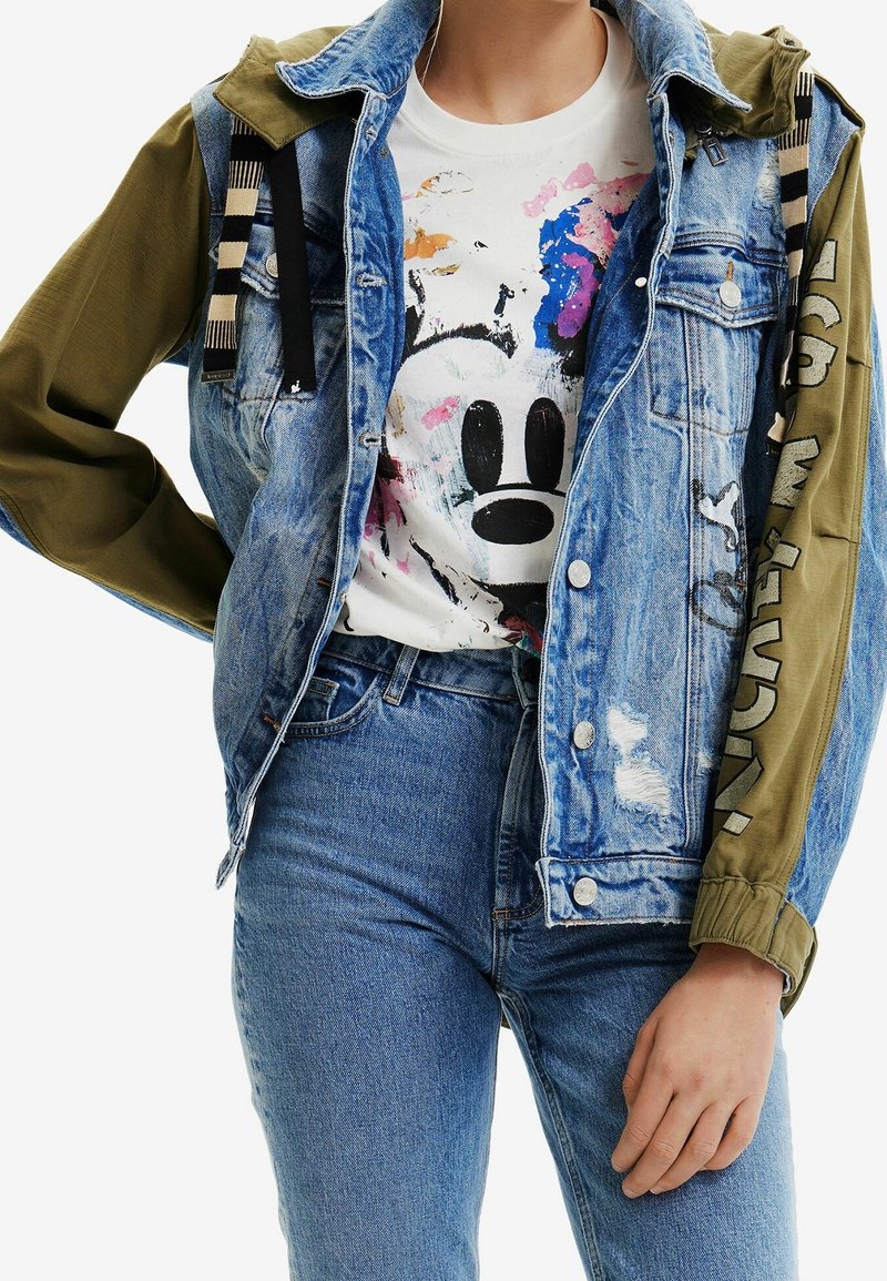 Chaqueta de denim con un acabado azul claro desgastado, mangas de color oliva y una camiseta gráfica que presenta un colorido diseño de Mickey Mouse debajo.