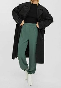 Przesadnie duży czarny trencz na dopasowanej czarnej bluzie, w połączeniu z zielonymi dresami z boczną kieszenią i elastycznymi mankietami, oraz białymi botkami za kostkę.