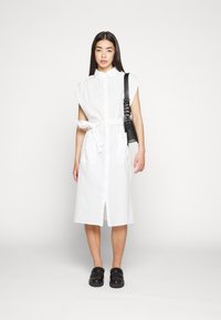 Vestido branco, sem mangas, com abertura frontal em botões e cinto. Tecido texturizado, até aos joelhos, com dois bolsos frontais. Usado com uma mala preta.