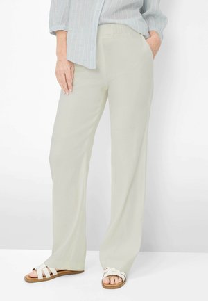 Person iført lyse beige bukser med vide ben, lyseblå stribet skjorte og hvide sandaler, stående med den ene hånd i lommen.