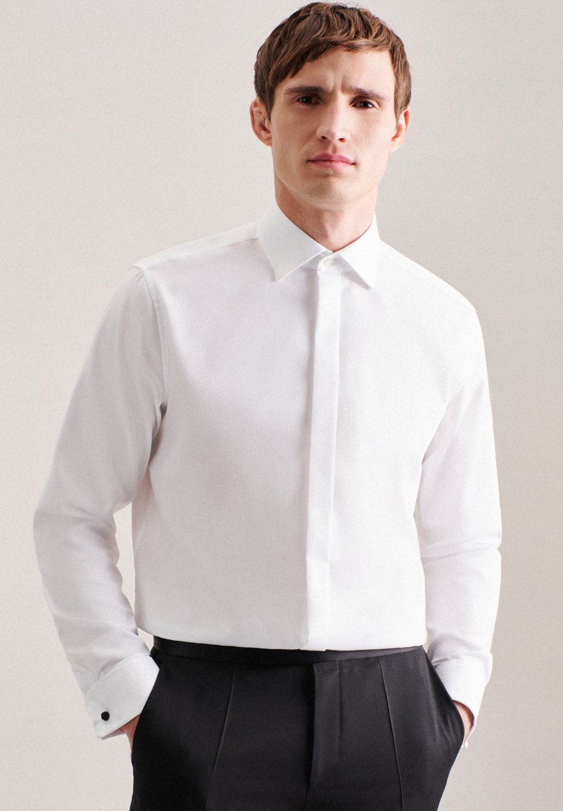 Camicia bianca con colletto classico e maniche lunghe, dotata di gemelli neri e una texture liscia e uniforme. Indossata con pantaloni formali neri.