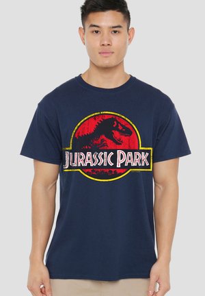 Man wearing navy blue Jurassic Park T-shirt with red and yellow logo, standing against plain light gray background.
