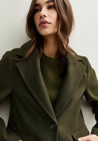 Blazer in misto lana verde oliva con rever a punta, doppi bottoni e spalle strutturate. Abbinato a un maglione verde a trama sottostante.