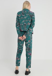 Ensemble costume floral bleu sarcelle comprenant une veste courte et un pantalon fuselé avec une texture brillante. Motif floral décoratif dans des tons de violet et de rose.