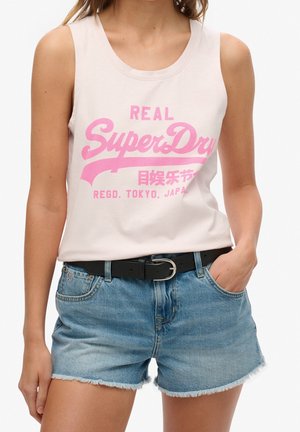 Kobieta nosząca biały bezrękawnik z różowym napisem "SuperDry" i niebieskie dżinsowe szorty z czarnym paskiem, jedna ręka w kieszeni.