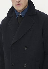 Jūras zila vilnas peacoat ar lieliem atlociem, divu rindu pogām un gludu apdari, uzvilkts pār tumšu džemperi un džinsu krekls.