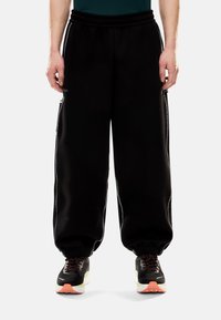 Pantaloni della tuta neri con vestibilità rilassata, vita elastica, tasche laterali con zip e polsini a coste. Presentano un dettaglio in piping bianco a contrasto sui lati.