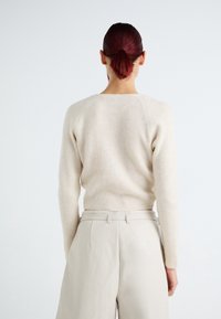 Femme aux cheveux rouge foncé attachés en queue de cheval, portant un pull côtelé beige et un pantalon taille haute crème, vue de dos sur un fond blanc.