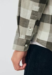 Szara i biała koszula w kratę z zapięciem na guzik, wykonana z miękkiego materiału, z widocznymi szwami i gładką teksturą. Noszona z ciemnymi spodniami sztruksowymi.