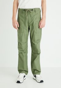 Olive green cargo pants with a drawstring waist, multiple front pockets, and a relaxed fit, paired with white and black sneakers.