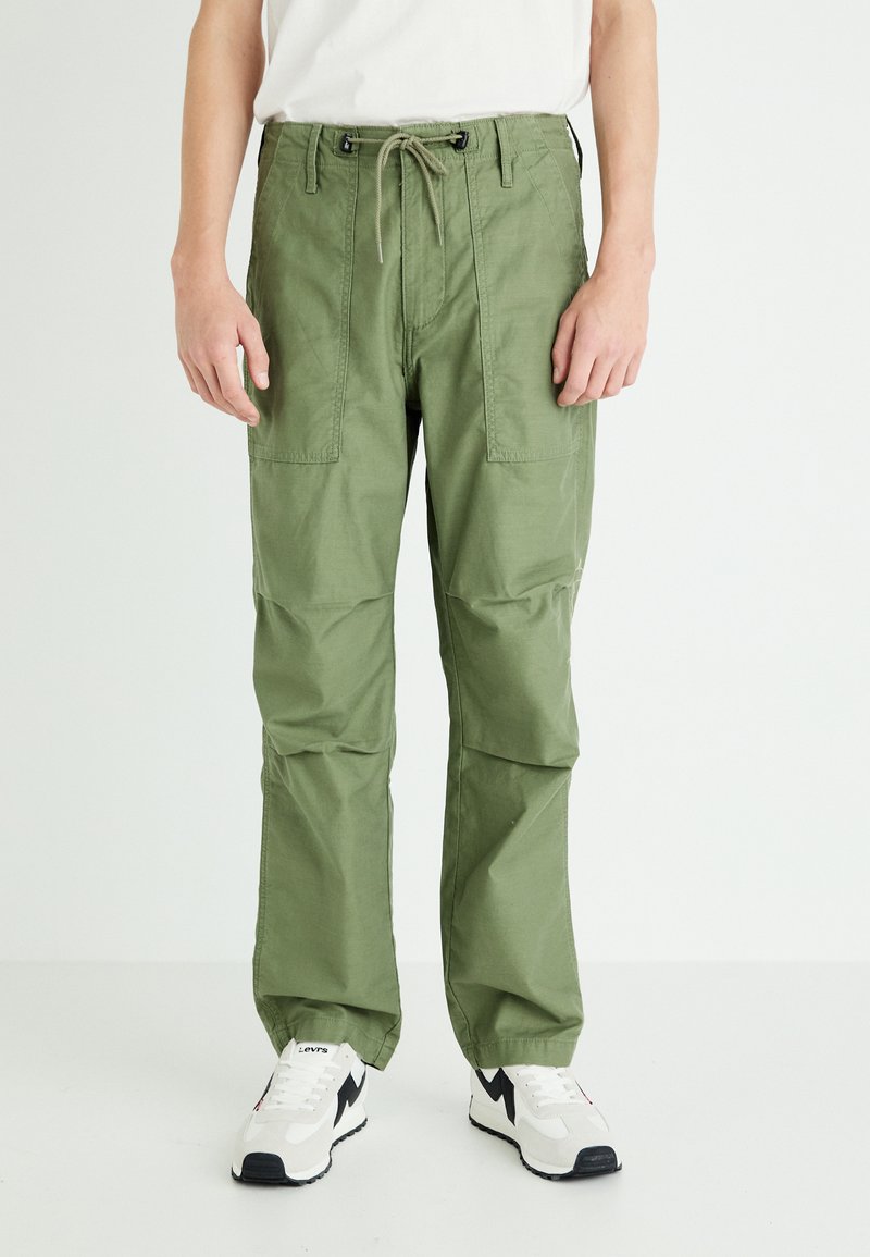 Olive green cargo pants with a drawstring waist, multiple front pockets, and a relaxed fit, paired with white and black sneakers.
