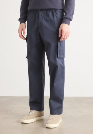 Homme portant un pantalon cargo bleu marine avec poches latérales et des baskets beiges, debout sur un sol clair devant un mur blanc.