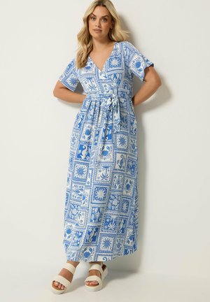 Femme portant une robe portefeuille bleue et blanche à motifs, manches courtes et sandales blanches, debout contre un mur clair uni.
