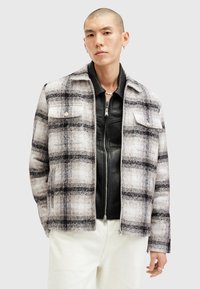 Ruudullinen takki off-white ja musta kangas, jossa on kaksi rintataskua ja kauluksellinen kaulus. Kerroksittain mustan vetoketjullisen nahkatakin päällä.