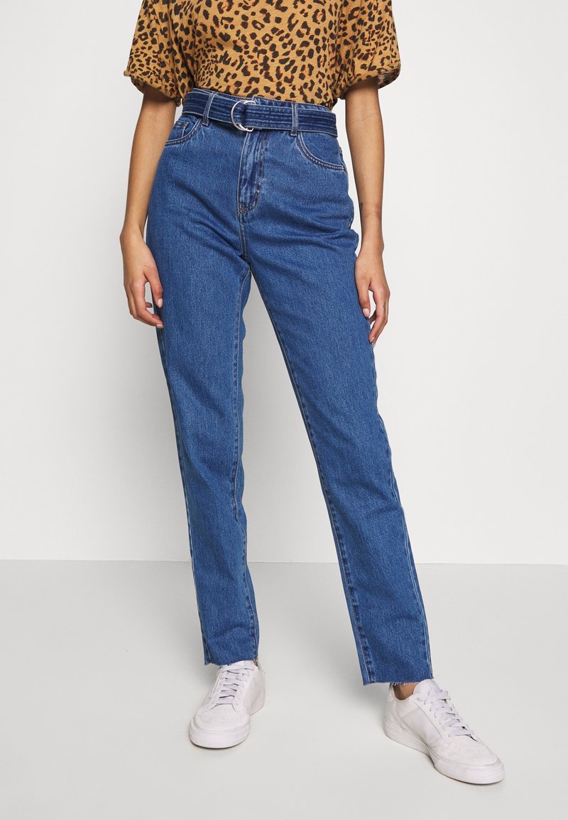 Personne portant un jean bleu taille haute avec une ceinture en denim, un haut à manches courtes imprimé léopard et des baskets blanches sur un fond uni.