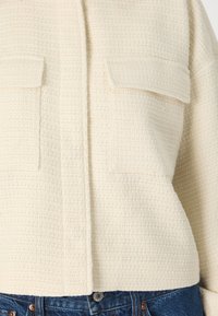 Krämfärgad, texturerad jacka med ett vävt mönster, som har två bröstfickor, knäppning framtill och en avslappnad passform tillsammans med blå denim.
