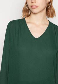 Mörkgrön långärmad blus med V-ringning, tillverkad av ett slätt tyg; har subtila veck vid axlarna och en minimalistisk design.