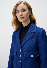 Blue textured jacket with a notched collar, two front pockets, and metallic buttons. Worn over a black turtleneck top.