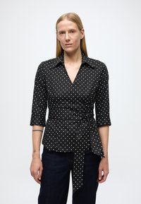 Zwarte wikkelblouse met witte stippen, driekwart mouwen, een puntige kraag en een ceintuurdetail in de taille. Gladde stof en een gestructureerd ontwerp.