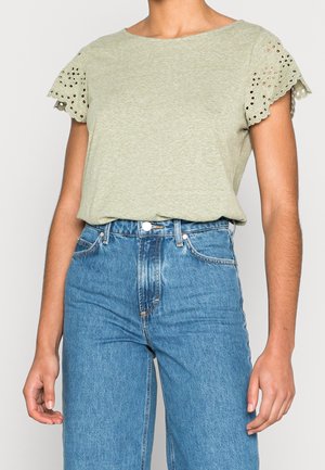 Persona vistiendo una camiseta de manga corta de color verde claro con detalles bordados de ojales en las mangas y jeans de mezclilla azul de talle alto.