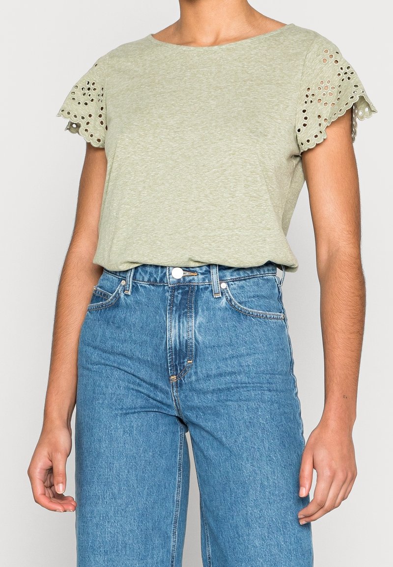 Personne portant un haut à manches courtes vert clair avec des détails brodés en œillets sur les manches et un jean en denim bleu taille haute.
