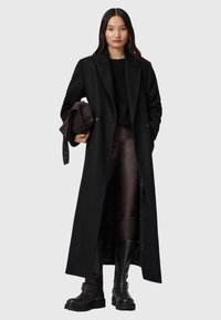 Czarny długi wełniany płaszcz z wyłamiem, podwójnie zapinany na guziki, noszony na ciemnym swetrze i brązowych skórzanych spodniach, w połączeniu z czarnymi botkami.