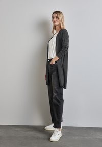 Vrouw staand met hand in de zak, gekleed in een wit shirt, een lange donkere cardigan, zwarte jeans en witte sneakers tegen een effen achtergrond.