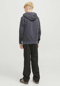 Felpa di colore grigio scuro con un ampio cappuccio, polsini e orlo a coste, abbinata a pantaloni neri e scarpe marroni. La vista è da dietro.