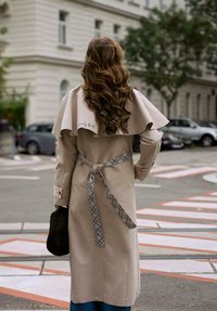 Žena s dlouhými kudrnatými vlasy, oblečená v béžovém trenčkotě s vzorovaným páskem, držící černou tašku, stojí na městské ulici poblíž zaparkovaných aut.