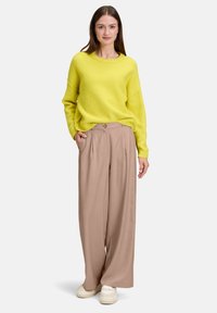 Kobieta z długimi brązowymi włosami, ubrana w żółty sweter, beżowe szerokie spodnie oraz białe trampki, stojąca z jedną ręką w kieszeni.