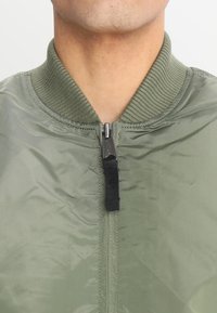 Olivgrön bomberjacka med ribbad krage och dragkedjestängning som har ett svart draghandtag. Slät, blankt tyg med minimala sömmar.