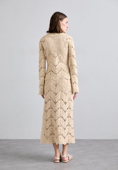 Beige lace dress with long sleeves, featuring a textured zigzag pattern and a tailored silhouette, reaching mid-calf with a side slit.