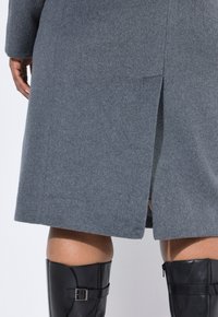 Cappotto grigio in lana con spacco posteriore, maniche lunghe e una texture morbida, abbinato a stivali neri lucidi fino al ginocchio con un'agenzia in metallo.