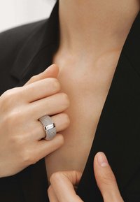 Anillo de malla plateada con una banda lisa y un acento rectangular. Lleva puesto en una mano contra una chaqueta negra, destacando su diseño texturizado.