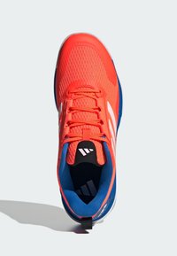 Scarpa sportiva rossa con una tomaia in rete traspirante, accenti blu, strisce bianche spesse e un pratico tassello sul tallone. Interno imbottito.