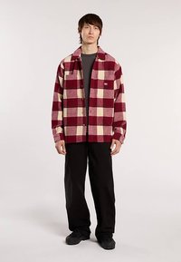 Camicia a quadri rossa e crema con maniche lunghe, bottoni frontali, vestibilità ampia e taschino sul petto, abbinata a pantaloni neri e scarpe nere.