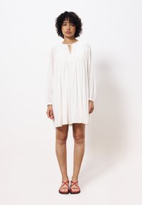 Weißes Langarmkleid mit gerafftem Ausschnitt, Schlitz-Bindungsdetail und strukturierter Stoffoberfläche. Kombiniert mit roten Riemchensandalen und bloßen Beinen.