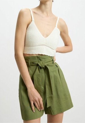 Femme portant un crop top en crochet blanc et un short taille haute vert olive avec une ceinture en tissu nouée, debout avec une main sur la hanche.