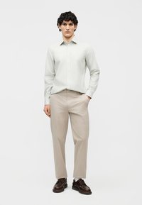 Mladý muž v svetlosivej pruhovanej košeli, béžových nohaviciach a tmavohnedých spoločenských topánkach stojaci s jednou rukou v vrecku.