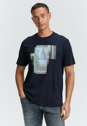 Camiseta de algodón azul marino con un diseño gráfico en capas en verde y gris, con el texto "PULSA >>> JUGAR TOM TAILOR" en el centro.