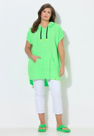 Frau steht in hellgrüner Kapuzen-Tunika, weißen kurzen Hosen und passenden grünen Sandalen vor schlichtem weißem Hintergrund.