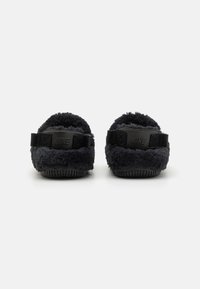 Svarta fluffiga tofflor med en mjuk, texturerad ovandel. Har ett justerbart svart spänne med präglad logotyp och en gummisula för bra grepp.