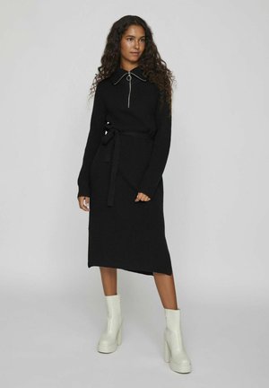 Vestido negro de punto con mangas largas, un cuello y una cremallera frontal. Cuenta con un lazo en la cintura y un dobladillo ligeramente asimétrico. Un par de botas de tacón blancas.