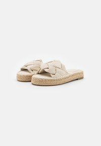 Sandales en tissu tissé de couleur beige clair avec un design de sangle tressée et une semelle en corde de jute. Plates et ouvertes à l'avant avec une texture douce.