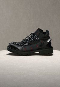 Botte en cuir montante noire avec semelle épaisse, lacets noirs et blancs à motifs, coutures rouges et texte blanc "DSQU" sur la bride du talon.