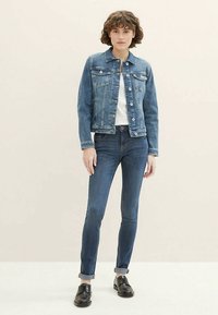 Jeans jakna srednje modre barve z gumbi spredaj, prsni žepi in rahlo skrajšanim dizajnom. Prilegajoče temne jeans hlače z zavihanimi hlačnicami in črnimi čevlji.