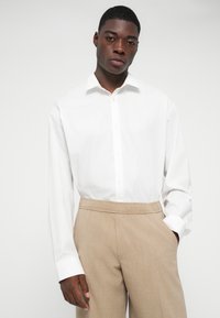 Weißes langärmliges Hemd mit Knöpfen, kombiniert mit hellbraunen, taillierten Hosen aus strukturiertem Stoff und elastischem Bund.