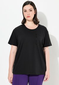 Musta t-paita lyhyillä hihoilla, pyöreällä kaula-aukolla ja vasemmalla puolella mustilla paljeteilla koristeltu tasku. Pehmeä, sileä kangas.